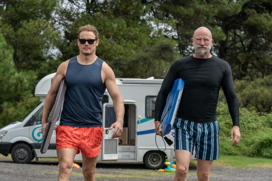 "men in kilts" temporada 2 - imagen promociona con Sam a la izquierda y Graham a la derecha, los dos con ropa de baño y una tabla de surf.