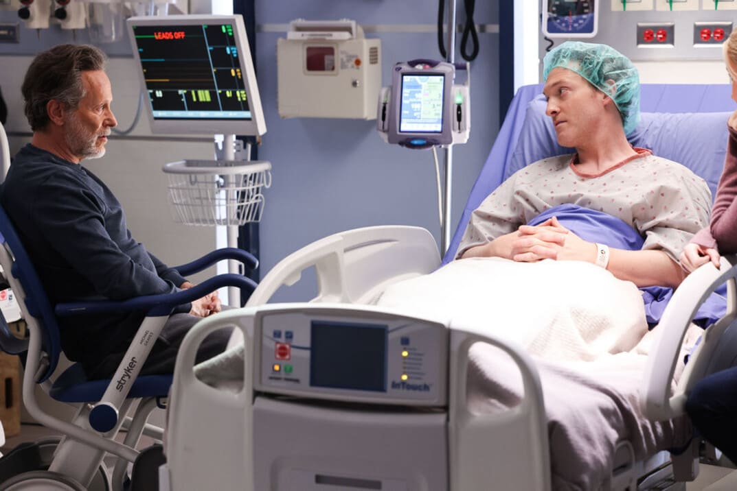 Imagen promocional de "Chicago Med" 9x03 Dean sentado en una silla de ruedas habla con su hijo Sean que está tumbado en la cama, listo para ir a quirófano